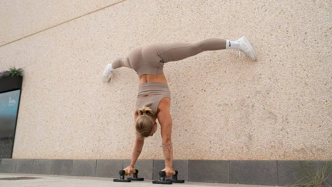 A woman performs a wall-supported straddle handstand on parallettes, wearing a beige workout outfit, against a textured wall.