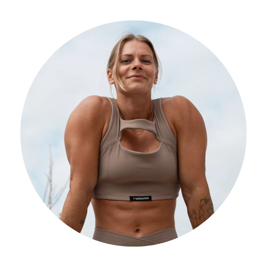A woman smiles confidently while holding a shrug position, wearing a taupe workout top, with a bright sky in the background.